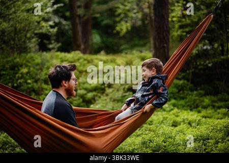 Ein fröhlicher Vater und Sohn teilen einen verspielten Moment in einer orangen Hängematte umgeben von Waldgrün, lächeln und genießen die gemeinsame Zeit. Stockfoto