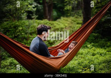 Ein fröhlicher Vater und Sohn teilen einen verspielten Moment in einer orangen Hängematte umgeben von Waldgrün, lächeln und genießen die gemeinsame Zeit. Stockfoto