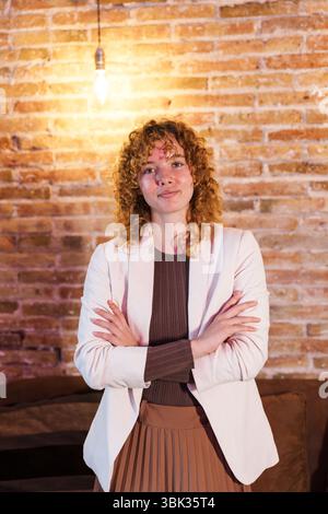 Selbstbewusste kaukasische Geschäftsfrau posiert mit überkreuzten Armen vor einer Ziegelmauer Stockfoto