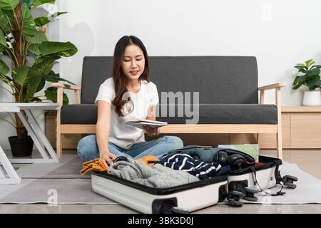 Reisevorbereitung. Eine Frau sitzt auf dem Boden und überprüft ihre Packliste, während sie ihren Koffer organisiert. Stockfoto