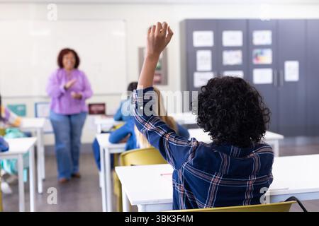 Schüler heben Hand im Klassenzimmer, während Lehrerin mit verschiedenen Schülern in der Schule interagiert Stockfoto