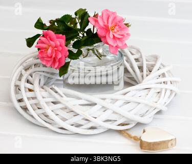 Rosen in einer kleinen Glasvase und ein weiß gewebter Weidenkranz mit Keramikherz Stockfoto
