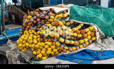 Verwitterte gelbe und braune Fischwagen und verworrene Netze stapelten sich auf einem Boot am Hafen, mit Teilen des Schiffes und Hafenszene im Hintergrund. Stockfoto
