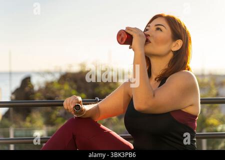 Eine junge Frau in Sportbekleidung ruht sich aus und rehydriert, während sie während einer Pause von ihrem Outdoor-Fitnesstraining aus einer wiederverwendbaren Wasserflasche trinkt Stockfoto