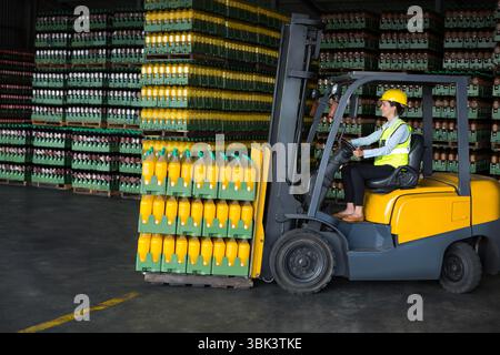 Der Gabelstaplerfahrer trägt Helm, die Warnweste hebt die Palette mit Orangensaftkisten im Lager an Stockfoto