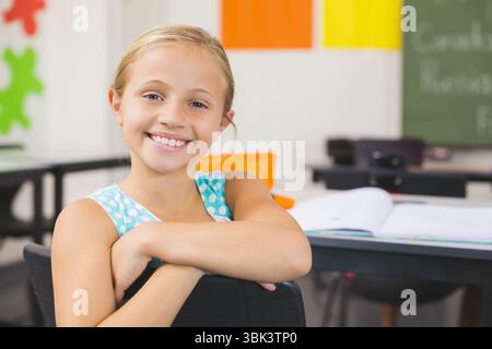 Lächelnde Schülerin lehnt sich im Klassenzimmer auf den Stuhl, mit Puzzledekorationen und Arbeitsbuch Stockfoto