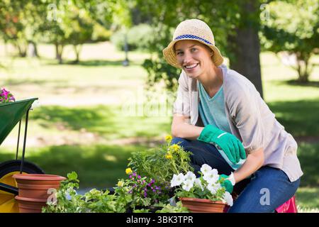 Frau kniet im sonnigen Garten mit Strohhut, pflanzt Blumen mit Schubkarre, Kopierraum Stockfoto