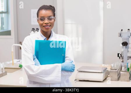 Afroamerikanerin mit Labormantel und Handschuhen, die blaues Bindemittel im Labor mit Mikroskopen hält Stockfoto