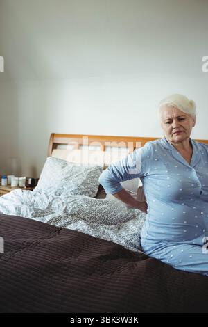 Ältere Frau, die im Schlafzimmer auf dem Bett sitzt und einen Pyjama trägt, der hinten am Nachttisch, Kopierraum gestützt wird Stockfoto