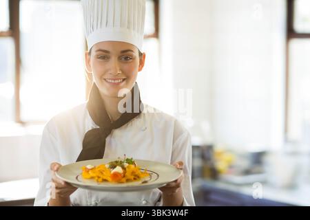 Weibliche Küchenchefin mit Plisseehut, die in der hellen Küche Nudelteller mit Kräutern und Käse hält Stockfoto