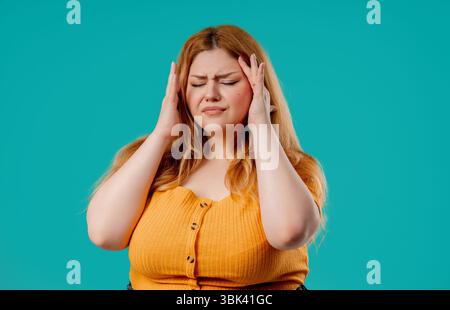 Frau in Übergröße, die Schläfe berührt, Kopfschmerzen hat, blaues Studio. Schmerzen. Stockfoto