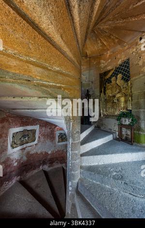 Die steinerne Wendeltreppe im Seitenturm des Château de Boussac, Département Creuse, Nouvelle-Aquitaine, Frankreich, windet sich um ein solides Wendeltreppenpfosten. Gerahmte Tapisseriefragmente auf hellem Stoff hängen an der rot verputzten Wand. Oben steht hinter einem Gitter ein vergoldetes Relief mit sitzenden Figuren, davor eine Laterne auf einem Sockel und eine Pflanze auf einem kleinen Holztisch. Die grauen Steinstufen sind abgenutzt, und die Steingewölbe tragen Spuren von gelblicher Farbe. Selbst die Wände im Treppenhaus sind mit Tapisserien geschmückt. Das Schloss ist eine mittelalterliche Festung mit einer reichen Geschichte. Marschall Jean de Brosse, ein c Stockfoto