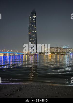 Nächtlicher Blick auf das Wasser in Dubai mit einer Brücke auf der linken Seite und dem beleuchteten Versace Hotel und dem D1 Tower auf der anderen Seite des Wassers Stockfoto