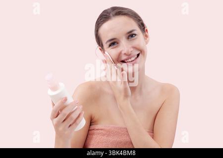 Lächelnde junge Frau, die ihr Gesicht mit Wattepad und Mizellenwasser reinigt. Hautpflege Routine, Make-up entfernen, frische gesunde Haut. Isoliert auf rosa Rückseite Stockfoto