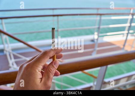 Lissabon, Portugal - 10. Juni 2025: Eine Hand hält eine Zigarette auf dem Deck eines Schiffes - das Bild steht für Rauchen an Bord, Ruhe auf See und Umgang mit dem Tabakkonsum während einer Fahrt und der Brandgefahr auf See. *** Eine Hand hält eine Zigarette auf dem Deck eines Schiffs das Bild steht für Rauchen an Bord, Pause auf See und den Umgang mit Tabakkonsum während einer Schifffahrt und die Gefahr durch Feuer auf dem Meer. Stockfoto
