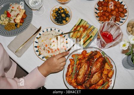 Nahaufnahme der Hände einer schwarzen Frau, die am Esstisch sitzt, mit verschiedenen festlichen Gerichten, die Reis, gegrilltes Gemüse und verschiedene Fleischgerichte genießen, Kamera-Blitz Stockfoto