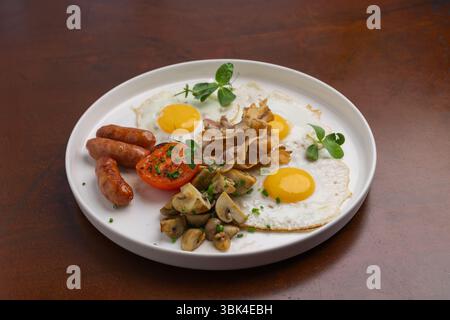 Ein köstliches Frühstück mit Spiegeleiern, Würstchen, gegrillten Tomaten und sautierten Pilzen, garniert mit Kräutern, serviert auf einem weißen Teller. Stockfoto