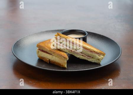 Herzhaftes Sandwich mit Schichten Fleisch und Käse auf einem runden schwarzen Teller neben einer kleinen Schüssel Dip-Sauce. Stockfoto