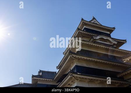 Der Turm der Burg Matsumoto in Nagano mit mehrschichtigen Ziegeldächern und Holzpaneelen wird von der linken Seite durch Sonnenlicht beleuchtet, wobei die Linsen gegen den klaren Himmel leuchten Stockfoto