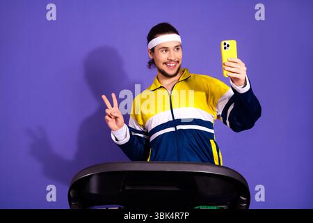 Aktiver athletischer Mann in Retro-Sportbekleidung lächelt und posiert mit einem Smartphone auf einem Laufband vor violettem Hintergrund Stockfoto