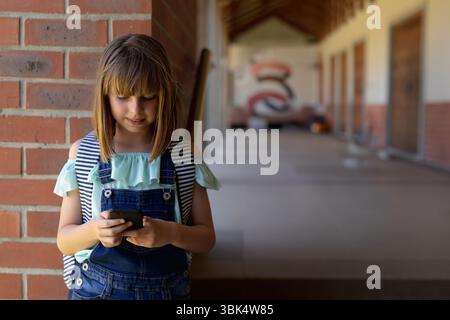 Weibliches Kind mit Smartphone auf Ziegelsäule im Schulflur mit Rucksack, Kopierraum Stockfoto