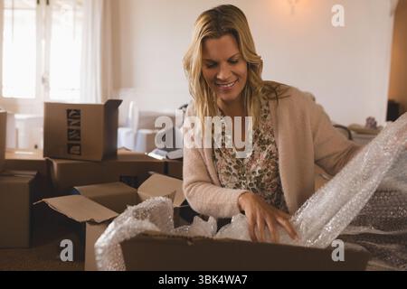 Erwachsene Frau, die am Tisch sitzt und Luftpolsterfolie aus offenen Kisten an einem durchsichtigen Fenster und Sofa zieht Stockfoto
