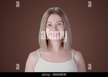 Porträt einer Frau mit glatten blonden Haaren, die lächelt und die Kamera vor braunem Hintergrund betrachtet Stockfoto