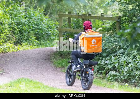 Rückansicht des Just Eat Liefermannes im Turban, der auf dem Weg mit einem Elektrofahrrad unterwegs ist, mit orangem Rucksack mit heißem Essen zum Mitnehmen zur britischen Lieferadresse rasst. Stockfoto