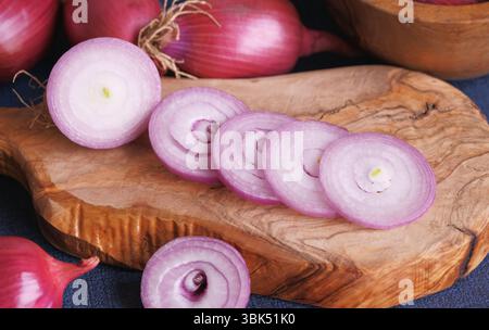 Tropea rote Zwiebelscheiben auf einem hölzernen Schneidebrett und ganze rote Zwiebeln im Hintergrund Nahansicht. Stockfoto