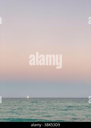 Ein ruhiges Segelboot gleitet über den Horizont unter einem pastellfarbenen Himmel aus zartem Rosa und Blau, der sich über ruhige türkisfarbene Meereswellen spiegelt. Stockfoto