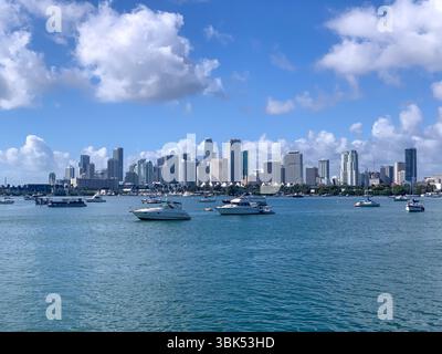 Ein heller, sonniger Blick auf die Skyline von Miami mit Booten, die auf ruhigem türkisfarbenem Wasser in der Biscayne Bay ankern. Hohe moderne Wolkenkratzer und flauschige Wolken Stockfoto