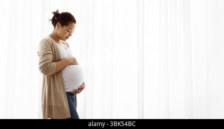Porträt Der Schönen Lächelnden Schwarzen Schwangeren Frau, Die Zärtlich Den Bauch Umschließt, Während Sie Vor Dem Fenster Zu Hause Steht, Glückliche Erwartungsvolle Mutter, Die Sie Genießt Stockfoto