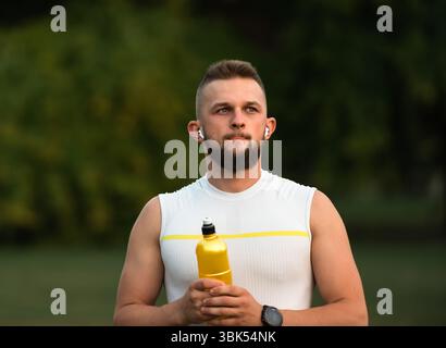 Ein junger, fitter Mann hört Musik über kabellose Kopfhörer und entspannt sich nach einem Lauf oder Workout in einem Sommerpark und zeigt einen aktiven, gesunden Lebensstil. Stockfoto