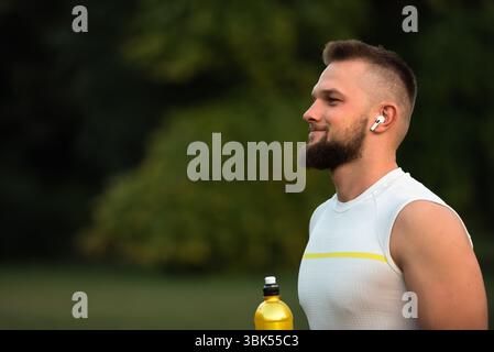 Ein junger, fitter Mann hört Musik über kabellose Kopfhörer und entspannt sich nach einem Lauf oder Workout in einem Sommerpark und zeigt einen aktiven, gesunden Lebensstil. Stockfoto