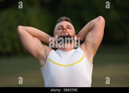Ein junger, fitter Mann, der sich vor dem Laufen oder Workout in einem Park dehnt, hört Musik über kabellose Kopfhörer mit geschlossenen Augen, um motiviert zu bleiben. Aktiv Stockfoto
