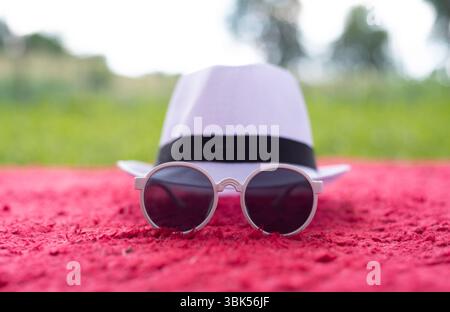 Ein modischer weißer Hut mit schwarzem Band und runder Sonnenbrille auf einer roten texturierten Decke. Sommer-Style, Urlaubsstimmung und trendige Accessoires Stockfoto
