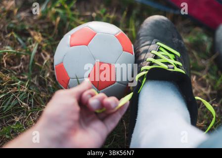 Eine Nahaufnahme einer Fußballszene im Freien mit einem leuchtenden orangen und weißen Fußball und einem schwarzen Fußballschuh mit hellgrünen Schnürsenkeln. Das Gras Stockfoto