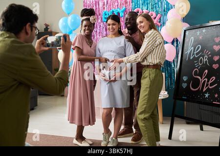 Freunde lächeln und berühren den Bauch der schwangeren Frau, während ein Mann sie mit dem Smartphone auf der Gender Reveal Party fotografiert Stockfoto