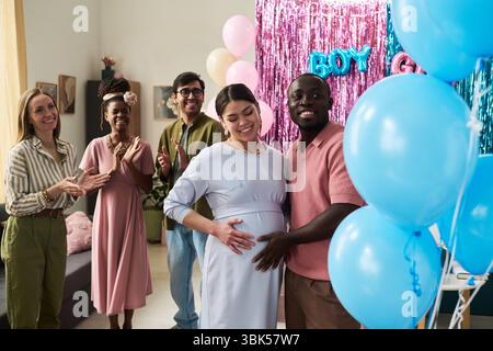 Schwangere asiatische Frau und Schwarzer Mann stehen zusammen lächelnd, während Freunde klatschen und bunte Ballons Hintergrund dekorieren Stockfoto
