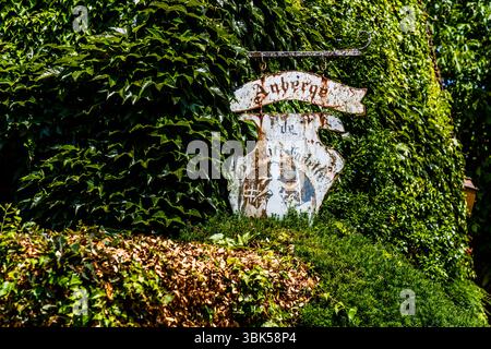 Ein von Efeu überwuchertes, verwittertes Schild der Auberge de la Petite Fadette hängt am Place Sainte-Anne in Nohant, Gemeinde Nohant-Vic, im Berry, Département Indre, Centre-Val de Loire, Frankreich, vor dem Anwesen. Das Metallschild besteht aus einer Schriftrolle mit der Aufschrift „Auberge“ und einem gebogenen Schild darunter. Rost hat die Schrift „de la Petite Fadette“ und eine verblasste Figur teilweise weggefressen. Das Schild hängt an einer Eisenstange vor einer Efeuwand, vor der ein Heck wächst. Die Gaststätte, ein Hotel und Restaurant, ist nach George Sands Roman „La Petite Fadette“ (Little Fadette) benannt fr Stockfoto