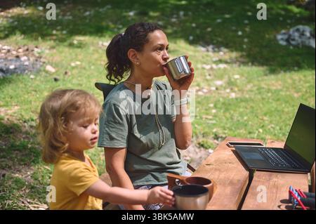 Eine Mutter genießt die Natur, während sie ferngesteuert an ihrem Laptop arbeitet, aus einem Becher trinkt, mit ihrem Kind in der Nähe. Die Szene zeigt die Work-Life-Balance in Stockfoto