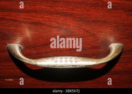 Ein Metallgriff, der an einer Holzschublade befestigt ist. Der Griff hat eine abgenutzte, leicht angetrübte Textur mit kleinen kreisförmigen Einbuchtungen in der Mitte. Stockfoto