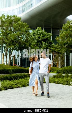 Ein Paar geht Hand in Hand, lächelnd und lachend, umgeben von üppigem Grün in einem städtischen Park an einem hellen, sonnigen Tag. Stockfoto