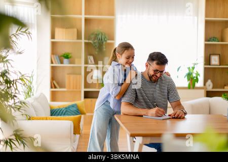 Ein Vater genießt eine gute Zeit mit seiner Tochter, während sie in einem schön gestalteten Wohnzimmer mit natürlichem Licht schreiben. Stockfoto
