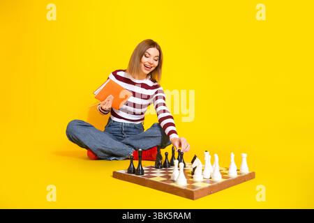 Junge Frau genießt Freizeit beim Schachspielen auf leuchtendem gelbem Hintergrund, lässigem Outfit und fröhlichem Ausdruck Stockfoto