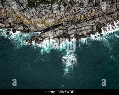 Aus der Vogelperspektive direkt über felsigen Klippen in raue Meere in einem verborgenen Küstenbild mit harten Granitfelsen Stockfoto