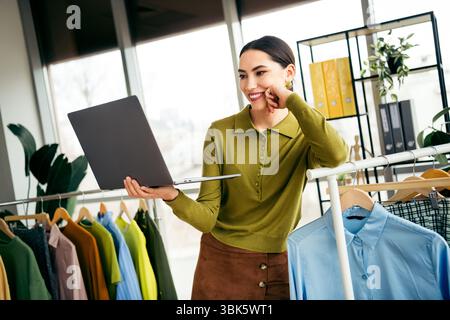 Attraktive Geschäftsfrau, die einen Ausstellungsraum in einem Atelier mit einem Laptop, umgeben von Kleiderständern in einem modernen Arbeitsbereich, verwaltet Stockfoto