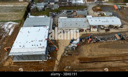 Arbeiter und schwere Maschinen sind auf einer Baustelle beschäftigt, auf der neue Geschäftsgebäude entstehen. Das Gebiet ist von Häusern und Feldern umgeben, was auf ein aktives Entwicklungsprojekt hinweist. Stockfoto