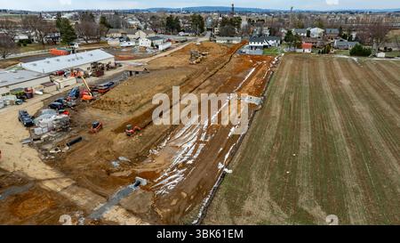 Bauprojekt Umgestaltung der Landschaft in der Nähe von Wohngebieten und Ackerland im frühen Frühjahr. Auf einer Baustelle in der Nähe von Häusern und offenen Feldern sind hochwertige photoschwere Maschinen im Einsatz. Der Boden wird geebnet und für die künftige Entwicklung vorbereitet, wobei der Kontrast zwischen städtischem und ländlichem Umfeld deutlich wird. Stockfoto