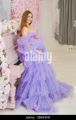 Schwangere Kaukasierin in fließendem Lavendelkleid posiert neben einem rosa Klavier. Der Hintergrund ist mit weichen Blumendekorationen versehen, die eine ruhige Atmosphäre schaffen Stockfoto
